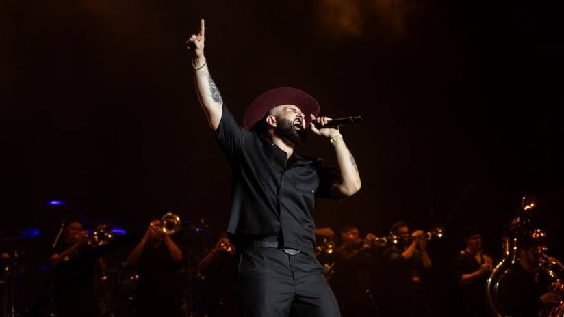 Singer performing on stage with a microphone while a brass band plays in the background.