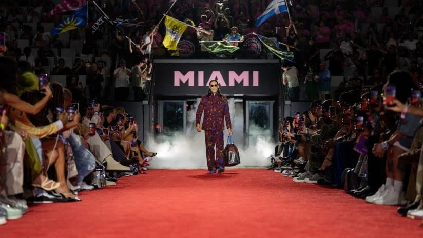 Model walking down a red runway as spectators watch and take photos during a Miami fashion show.